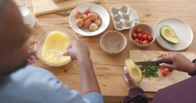 African couple starting meal prep, whisking eggs, chopping avocado and tomatoes at kitchen counter