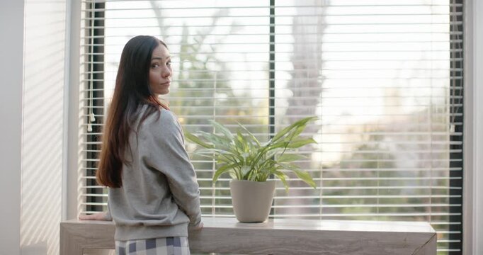 Asian woman looking out window, turning to camera, smiling at viewer by console plant in pajamas