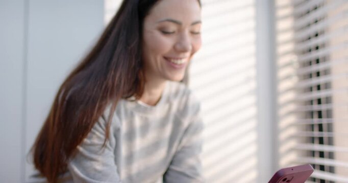 Adult woman tapping pink phone after ping scrolling smiling while reading text at counter by blinds