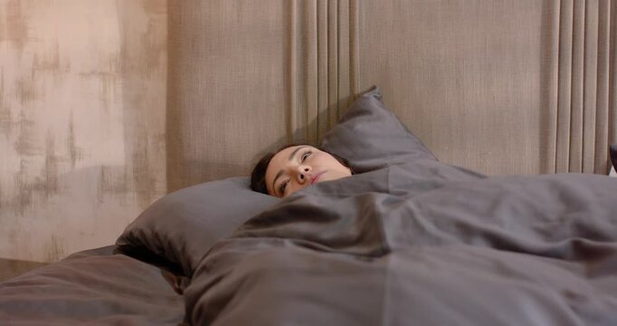 Adult female waking, lying in bed under gray duvet by paneled headboard, opening eyes, rubbing eyes