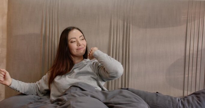 Adult female waking from sleep and stretching, sitting up on bed under grey duvet, smiling