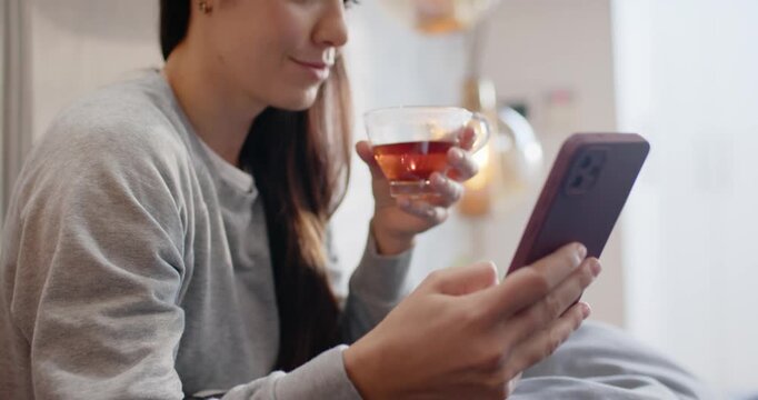 Woman holding smartphone with mauve case, reading messages and sipping tea on bed smiling