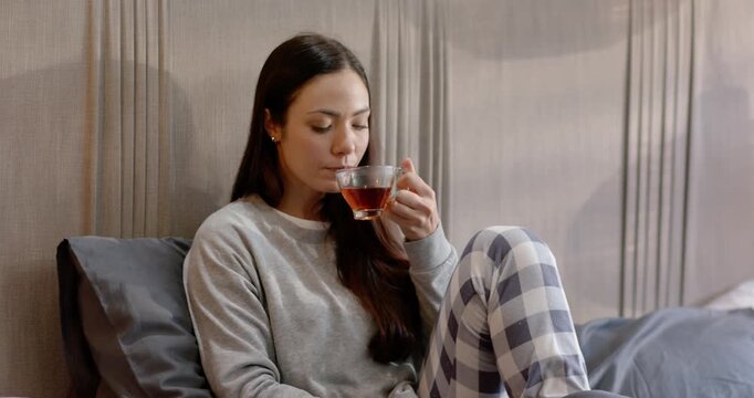 Woman lifting glass teacup to lips, sipping tea, relaxing on bed in gray sweatshirt and smiling