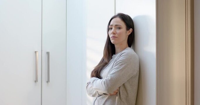 Adult woman 30s leaning by wall beside cabinets uncrossing, rubbing forehead from window light