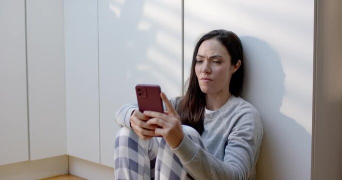 Woman in gray sweatshirt sitting on kitchen floor, scrolling pink phone after alert, calming