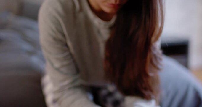 Woman in grey top and plaid pajamas sitting on dark couch, clasping hands, camera pulling back