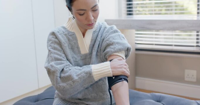 Asian woman sitting on ottoman at window rolling sleeve adjusting cuff and monitor for BP check