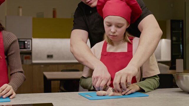 Unrecognizable male chef guiding Caucasian girl cutting raw chicken breast on cutting board. Video is suitable for promoting children culinary schools and educational cooking programs
