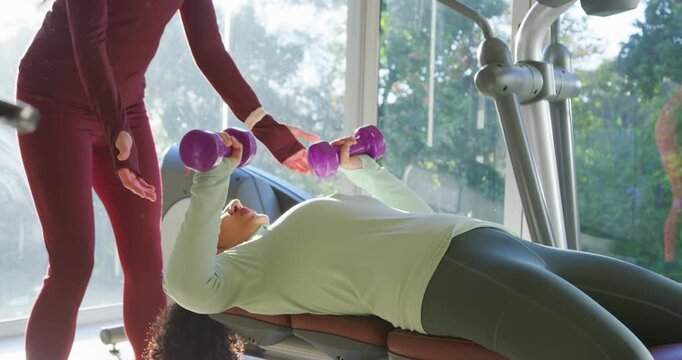 Female trainer cueing African American trainee pressing purple dumbbells at bench for safety