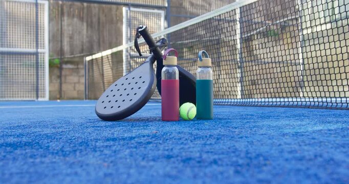 Padel racket and two reusable bottles are remaining on blue turf after match ends, ball resting