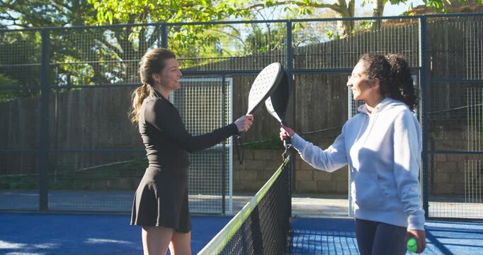 Female duo in sporty gear approaching net on padel court, tapping paddles with green ball pregame