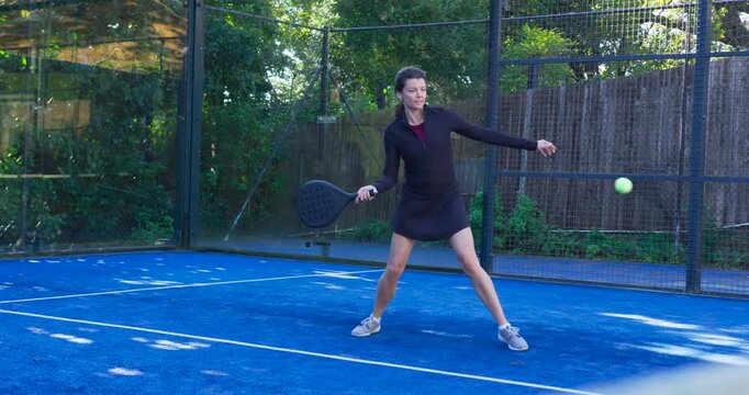 Adult woman hitting ball from right with paddle, returning shot on blue court wearing sportswear