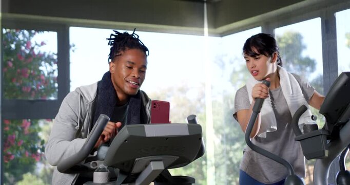 Diverse female friends working out in gym, placing red smartphone on bike console, sharing laughter