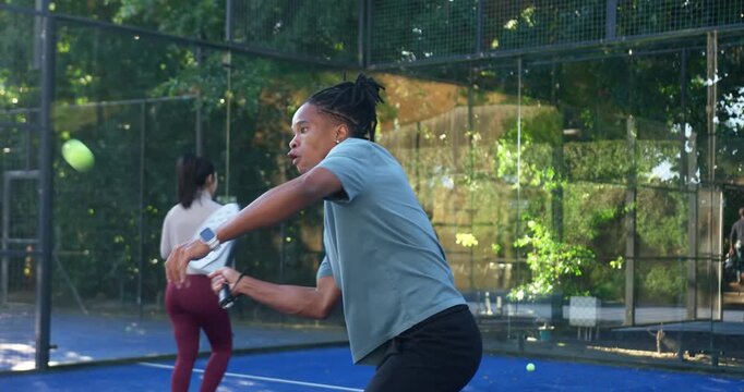 African American pair, male tracking ball, swinging forehand racket on blue padel court, returning