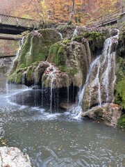 A serene waterfall cascades over moss-covered rocks in an autumn forest setting. © Letitia
