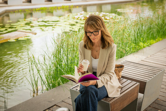 Confident blond haired woman reading a book with coffee during a relaxing break in a modern city park