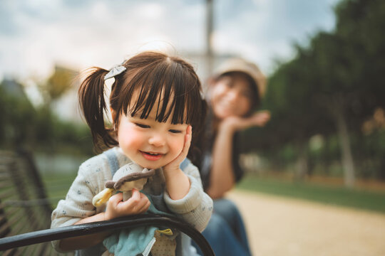 Cute little Asian girl smiling and resting chin on hand while sitting on park bench with her mother behind, happy toddler child holding a toy on outdoor family vacation for parenting and childhood