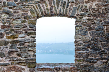 Obraz premium Ancient Stone Archway Framing a Misty Lakeside View. Weathered stone wall with an arched opening framing a calm lake and distant misty shoreline beneath an overcast sky.