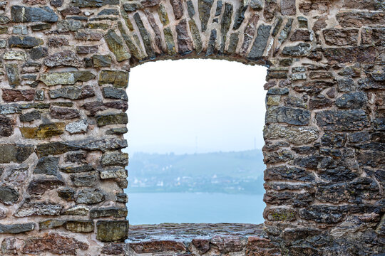 Ancient Stone Archway Framing a Misty Lakeside View. Weathered stone wall with an arched opening framing a calm lake and distant misty shoreline beneath an overcast sky.