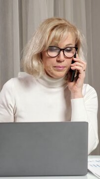 Focused mature businesswoman in glasses talking on a mobile phone, using a laptop, and working with documents at her desk, multitasking to solve a business problem in her modern office