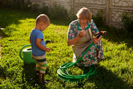 A grandmother shows her grandson how to use a watering hose to take care of plants in their garden while enjoying the warm afternoon sun