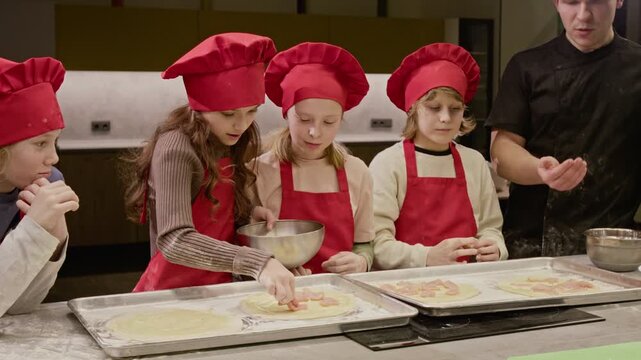 Caucasian children wearing chef hats preparing pizza during cooking class with male instructor. Video ideal for promoting culinary schools, children educational programs, or family activities