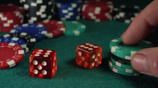Two colorful dice placed on a green felt surface with a hand reaching for a stack of teal poker chips in a casino setting