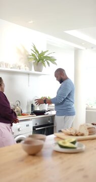 Vertical video: African couple preparing dinner in home kitchen, man tipping greens from clear bowl