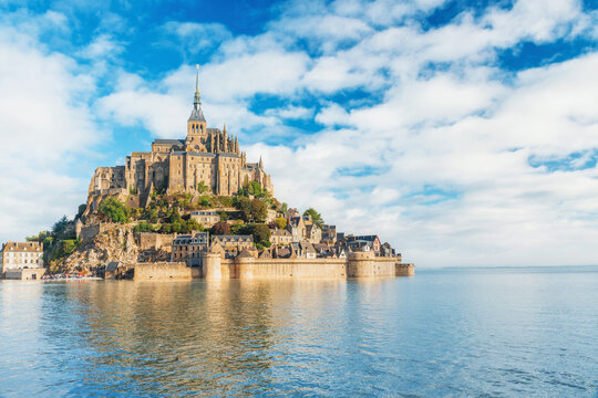 Mont Saint Michel abbey surrounded by calm high tide waters on cloudy day in Normandy, France. Tidal island with monastery in Normandie. Cultural tourism, coastal scenery and historic destination