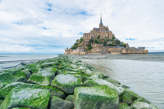 Mont Saint-Michel abbey above moss covered rocks under cloudy sky with scenic coastal landscape and medieval architecture in Normandy, France. Tidal island with monastery in Normandie