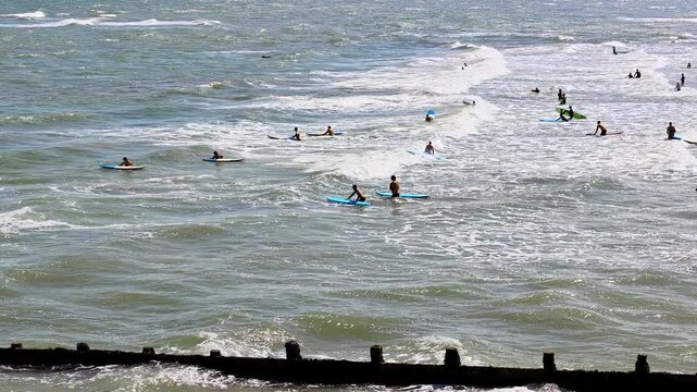 Static distant shot of a surf school session on Cromer Beach, North Norfolk coast. Surfers practise on the waves beneath a sunny sky, with a scenic seaside backdrop and coastal views.