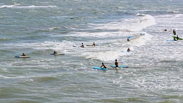 Panning shot of a distant surf school session on Cromer Beach, North Norfolk coast. The camera sweeps across surfers learning in the waves, with a scenic seaside backdrop under a bright sky.