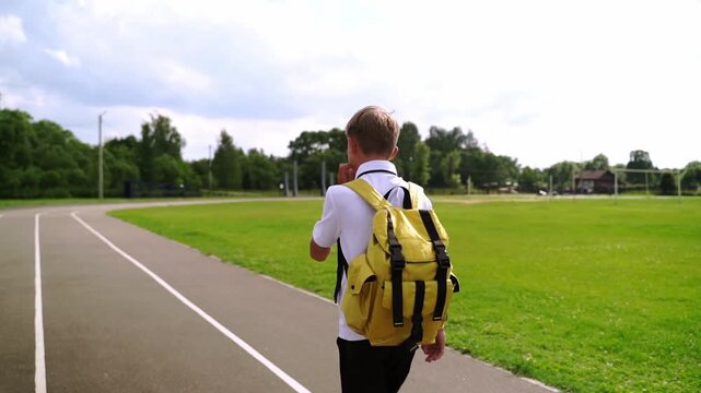 Boy walks away with yellow backpack on track. Green field and trees surround the path. He appears calm and focused. Perfect for school or outdoor activity themes. No animals or plants visible