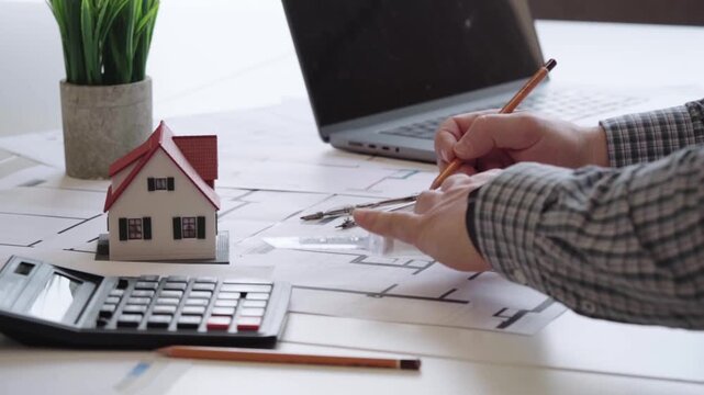 Hands sketch blueprints with precision and care. Small model home sits beside calculators and tools. Laptop, pencils, and compasses suggest detailed planning
