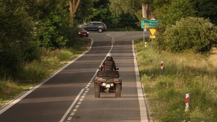 Zdjęcie przedstawia osobę jadącą quadem po asfaltowej drodze wśród zieleni. W oddali widać skrzyżowanie, znaki drogowe oraz zaparkowane samochody. © tomo81