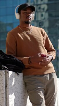 Vertical video: Leaning man in brown sweater and headphones, waiting on ledge holding red item