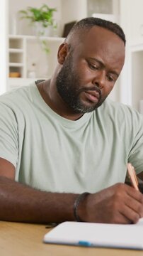 Vertical video: Picking pencil man in green tee writing in notebook at home for work checking phone