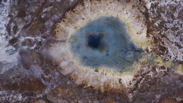 Aerial top down view of an Iceland Strokkur geyser pool, square dark blue vent, pale rings, yellow sulfur, white silica, rust brown ground, steam drift, bubbling, daylight