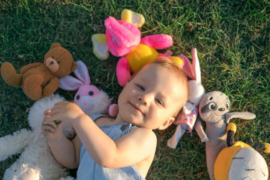 The baby is lying on the grass, surrounded by a pile of stuffed animals.