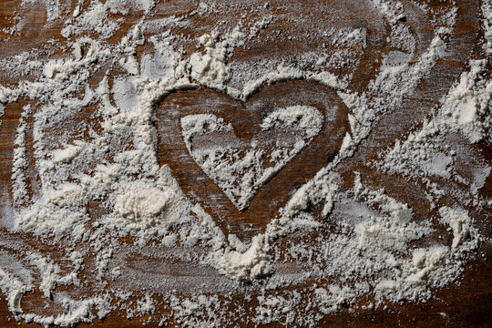Heart shape outline drawing in white flour on dark wooden baking background. White wheat flour scattered on rustic wooden table with hand drawn heart shape in center for baking and love concept