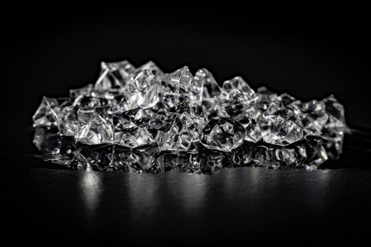 Pile of clear crushed ice cubes with reflection on black background, closeup. Heap of shiny transparent acrylic ice cubes reflecting on dark glossy surface for cold beverage drink concepts