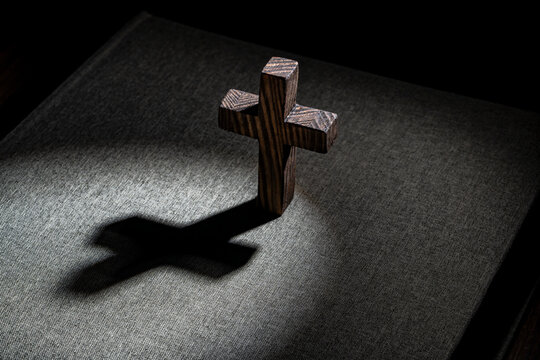 Wooden Christian cross casting long shadow on holy bible book cover, closeup. Small dark wooden cross stands on a fabric texture bible cover casting a strong dramatic shadow in selective spot light