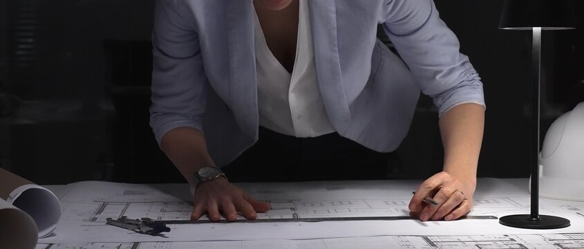 Female architect working on blueprints, measuring building plans at night, designing new construction project. Business and architect people concept