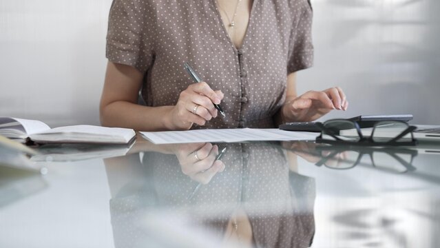 Businesswoman accountant, wearing brawn dotted blouse, is using a calculator to crunch numbers and jotting down meticulous notes, at sleek glass desk in a contemporary office. Taxes, audit in business