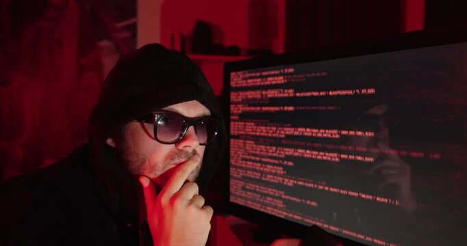 Hooded man looking at a computer screen filled with source code, representing hacking, cybersecurity threats, online crime, and digital security.