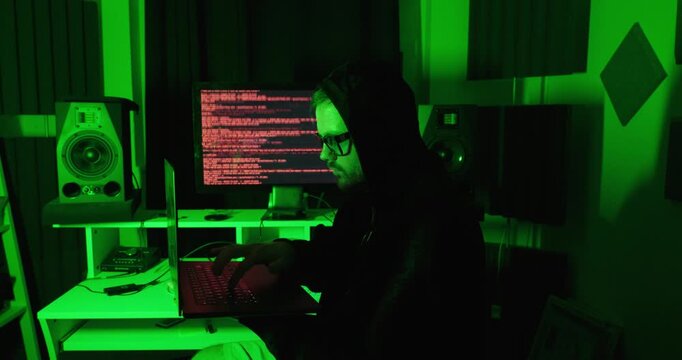 Hooded hacker working on a laptop in a dark room with computer code on the monitor, representing cybersecurity, hacking, cybercrime, and digital threats.