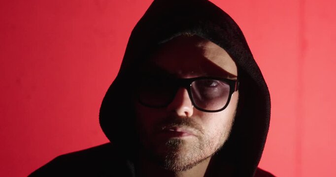 Close-up portrait of a hooded man wearing sunglasses against a red background, representing cybercrime, anonymity, hacking, and digital security threats.