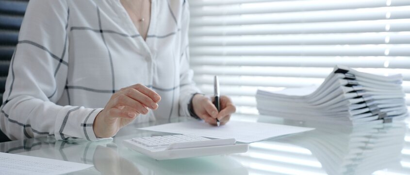 Woman's hands using a calculator and holding a pen, working with financial documents and a pile of papers on a clear desk in an office setup