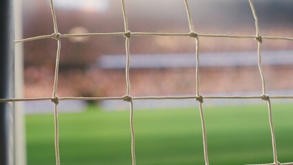 Empty goal net and post in soccer field viewed from behind rope, pitch with blurred crowd seating and bright arena light. Goal frame with selective focus, football gear, highlighting stadium crowd. © DC Studio