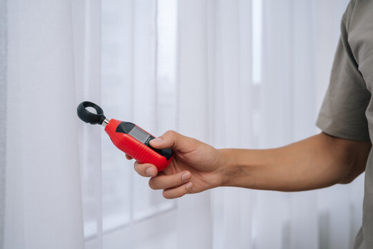 Close-up hand of unrecognizable man measuring natural light by window with digital luxmeter, testing illumination levels for optimal home brightness, energy efficiency and comfortable daylighting.
